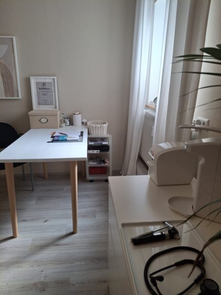 Helles Zimmer mit Tisch, Stuhl, Nähmaschine und Bücherregal in der Ecke.