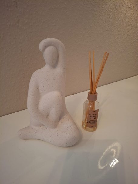 Sitzende Figur aus weißem Material neben einem Duftspender mit Stäbchen.