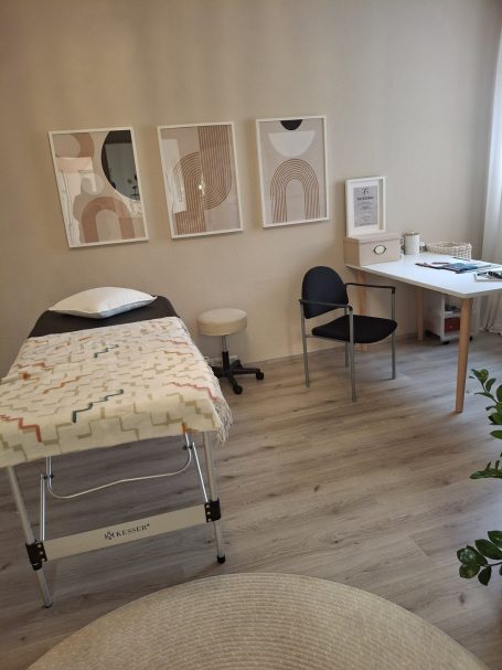 Behandlungsraum mit Liege, Stuhl, Tisch und Bildern an der Wand.