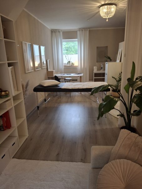 Ruhiger Raum mit Behandlungstisch, Pflanzen und großen Fenstern für Tageslicht.