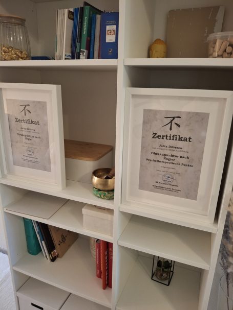 Zwei gerahmte Urkunden auf einem Regal, daneben Bücher und ein Behälter.